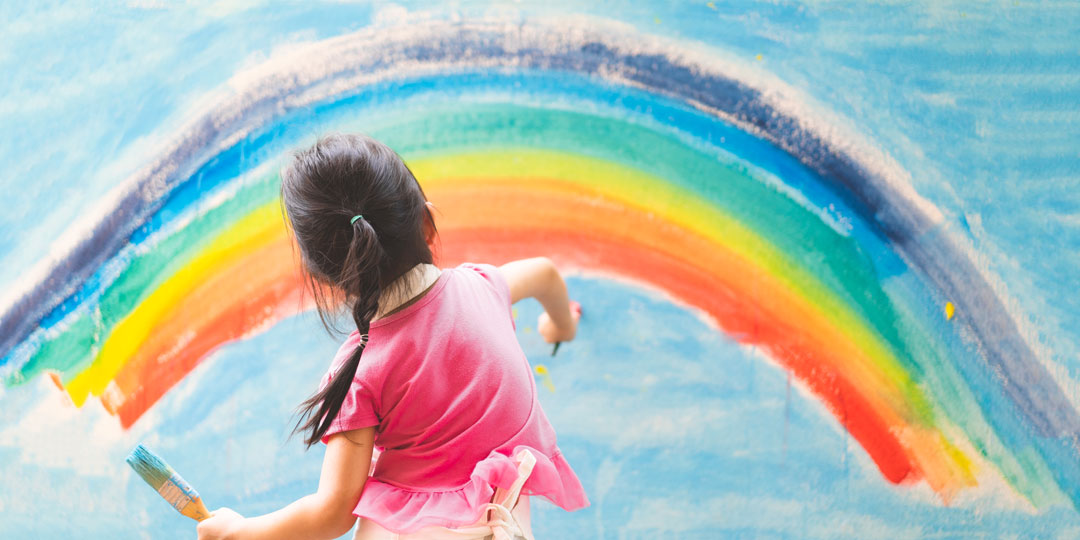 Flicka med stora penslar i händerna målar en regnbåge på en vägg.
