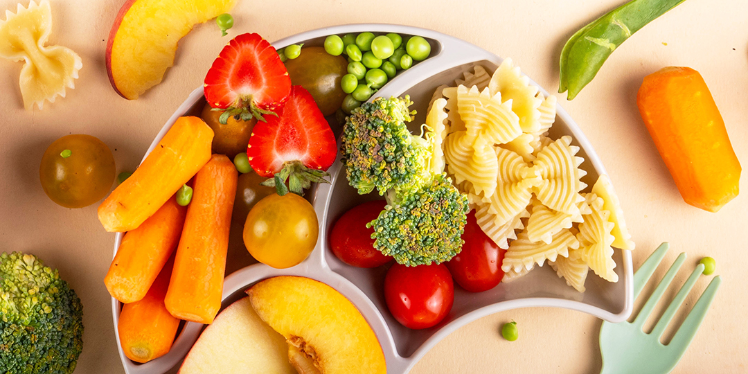 En mattallrik med grönsaker, pasta, frukt och bär bredvid en barngaffel.