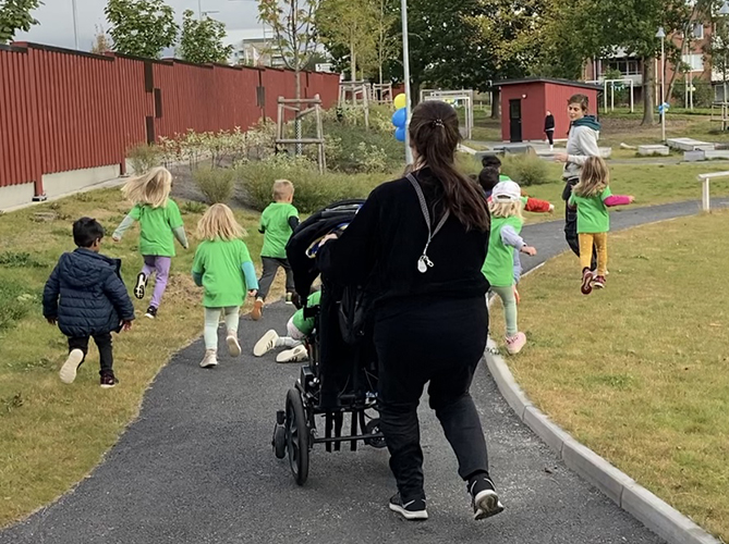 Förskolebarn som springer och pedagog som kör vagn ute på förskolegård.