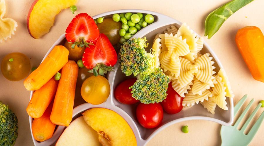 En mattallrik med grönsaker, pasta, frukt och bär bredvid en barngaffel.