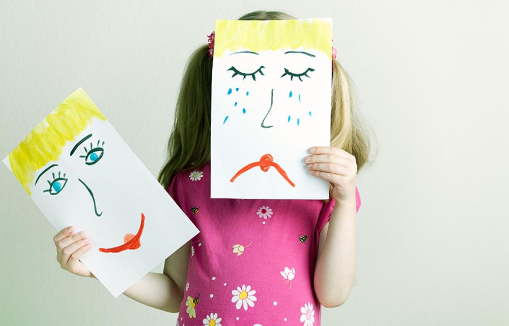 Ett förskolebarn håller upp två teckningar som illustrerar sorg och glädje