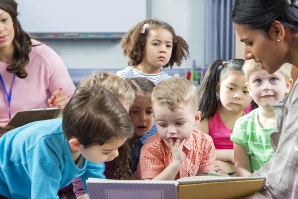 Bild på barn som sitter på golvet och tittar i en bok tillsammans med två pedagoger.