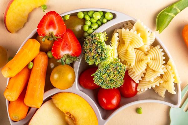 En mattallrik med grönsaker, pasta, frukt och bär bredvid en barngaffel.